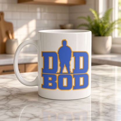 A white ceramic coffee mug with a blue and yellow 'DAD BOD' graphic, sitting on a marble kitchen counter with a blurred kitchen background.