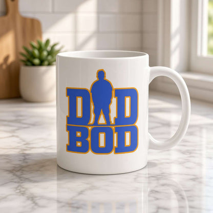A white ceramic coffee mug with a blue and yellow 'DAD BOD' graphic, sitting on a marble countertop in a bright kitchen setting.