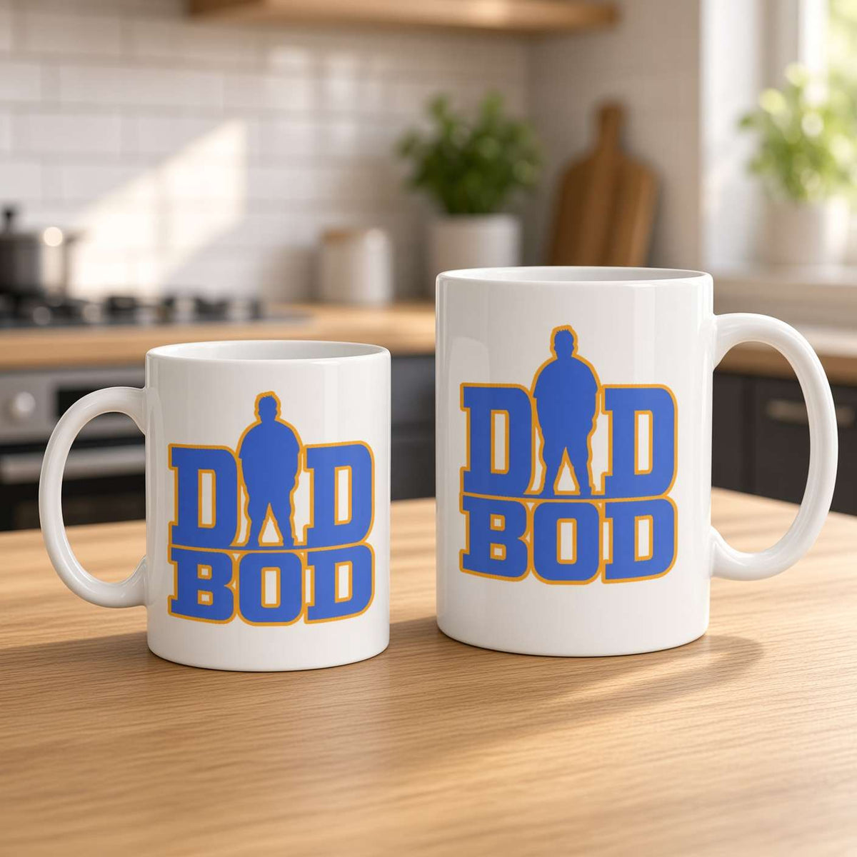 Two white ceramic mugs with a blue and yellow 'DAD BOD' graphic design, displayed on a wooden kitchen table with a blurred kitchen background