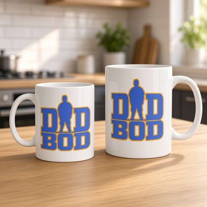 Two white ceramic mugs with a blue and yellow 'DAD BOD' graphic design, displayed on a wooden kitchen table with a blurred kitchen background