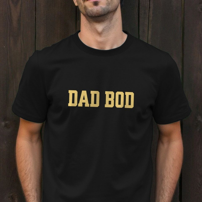 Person wearing a black funny slogan dad t-shirt with 'DAD BOD' slogan in yellow against a wooden background