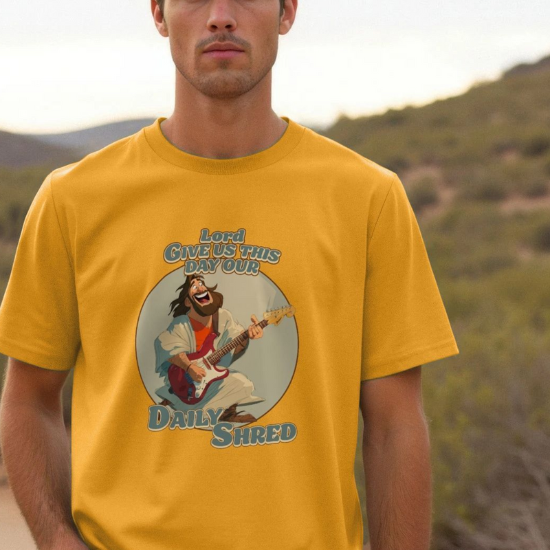 Man wearing a yellow t-shirt with a funny graphic design of a jesus playing guitar and text 'Lord Give Us This Day Our Daily Shred' against a natural background.