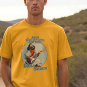 Man wearing a yellow t-shirt with a funny graphic design of a jesus playing guitar and text 'Lord Give Us This Day Our Daily Shred' against a natural background.