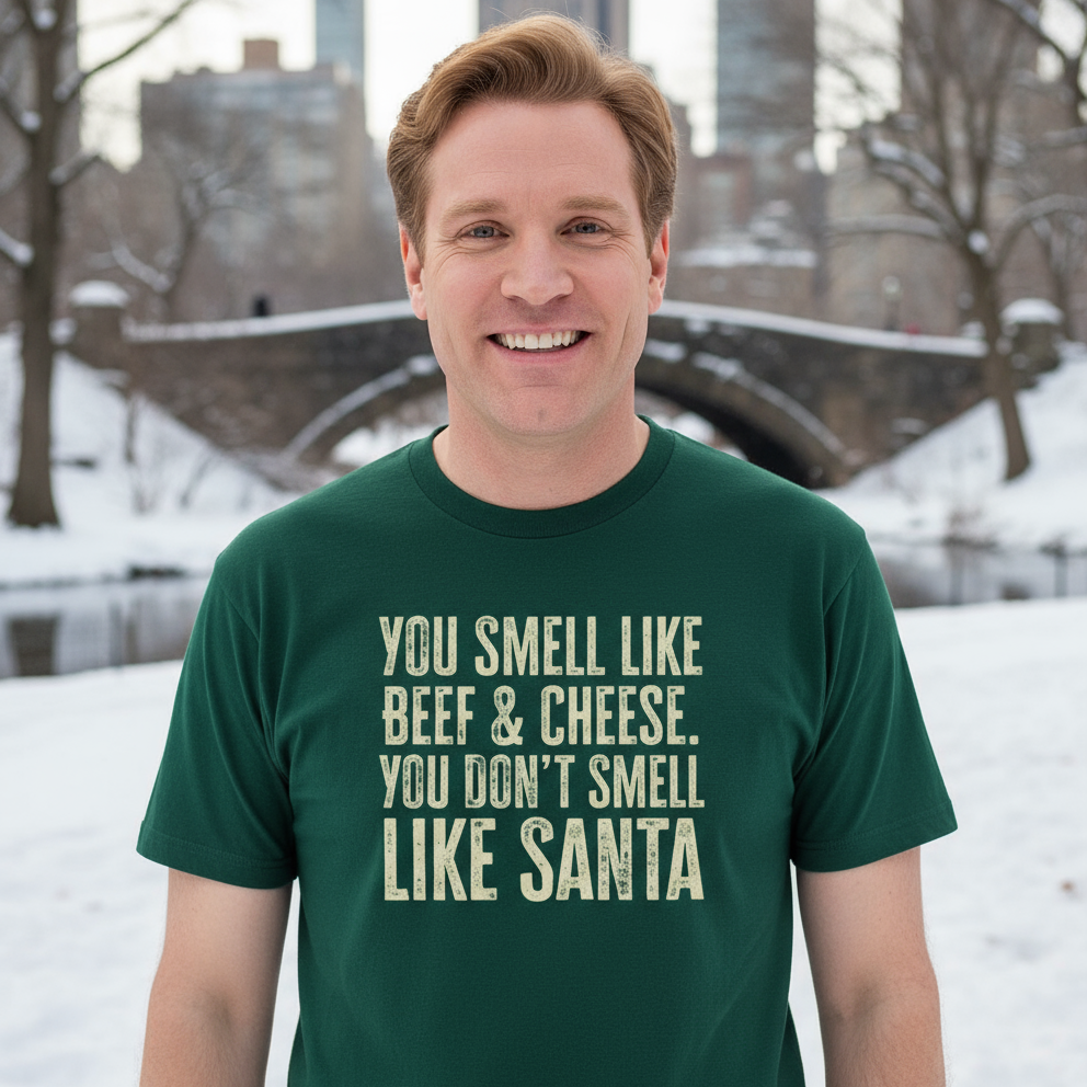 Man wearing a green Funny Christmas t-shirt with the humorous text "You smell like beef & cheese. You don't smell like santa" in a snowy New York setting