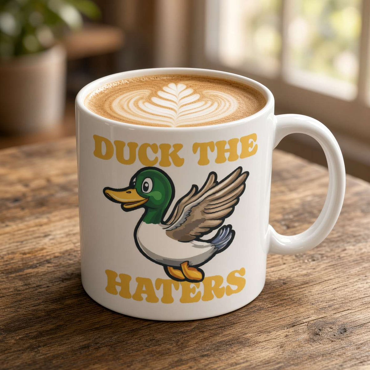 A white mug of latte with leaf-shaped foam art sits on a rustic wooden table. It features a cartoon duck and bold gold “Duck the Haters” text, creating a playful, upbeat mood.