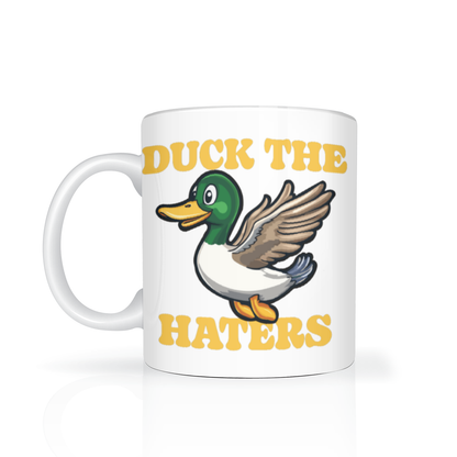 A white ceramic mug shows a cheerful cartoon mallard and bold yellow text reading “DUCK THE HATERS,” creating a playful, upbeat mood.