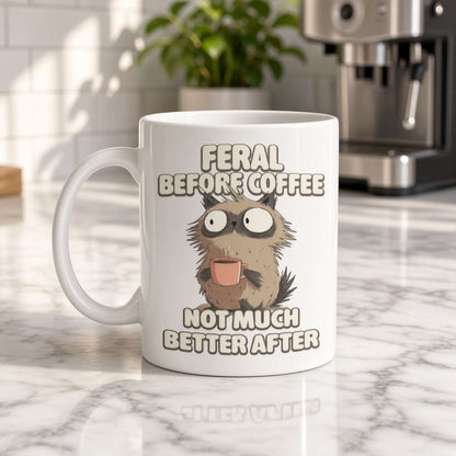 White ceramic coffee mug featuring a frazzled, wide-eyed cat holding a pink cup and the humorous text “Feral before coffee, not much better after.” Playful, relatable tone.