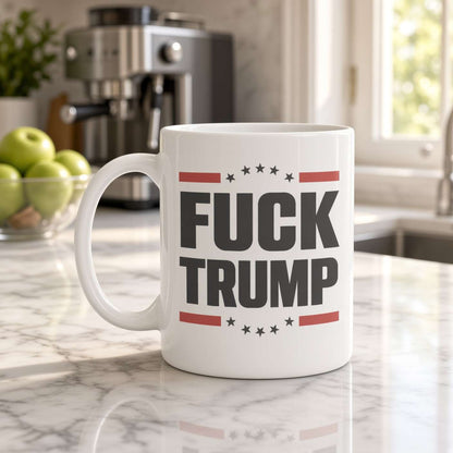 White “FUCK TRUMP” mug with red stripes and black stars on a marble kitchen counter near a coffee machine and green apples; a defiant political message.