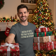 Man holding a gift with funny slogan on a funny Christmas T-shirt, surrounded by Christmas presents and decorations.