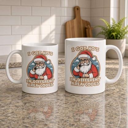 Two white coffee mugs on a kitchen counter feature a cartoon Santa and the phrase “I got hos in different area codes,” creating a playful, irreverent holiday mood.