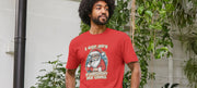 Man in a red funny Christmas t-shirt with humorous Santa print and text, stands in a lush indoor garden. The scene is lighthearted and festive.