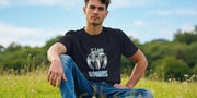 A man sits on grass in a field, wearing a black Funny Animal T-shirt with cartoon birds and 