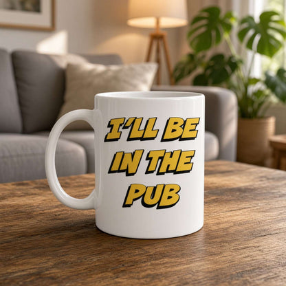 White ceramic mug reading “I’ll be in the pub” in bold yellow-and-black lettering sits on a wooden table, with a cozy sofa, lamp, and leafy plant behind it.