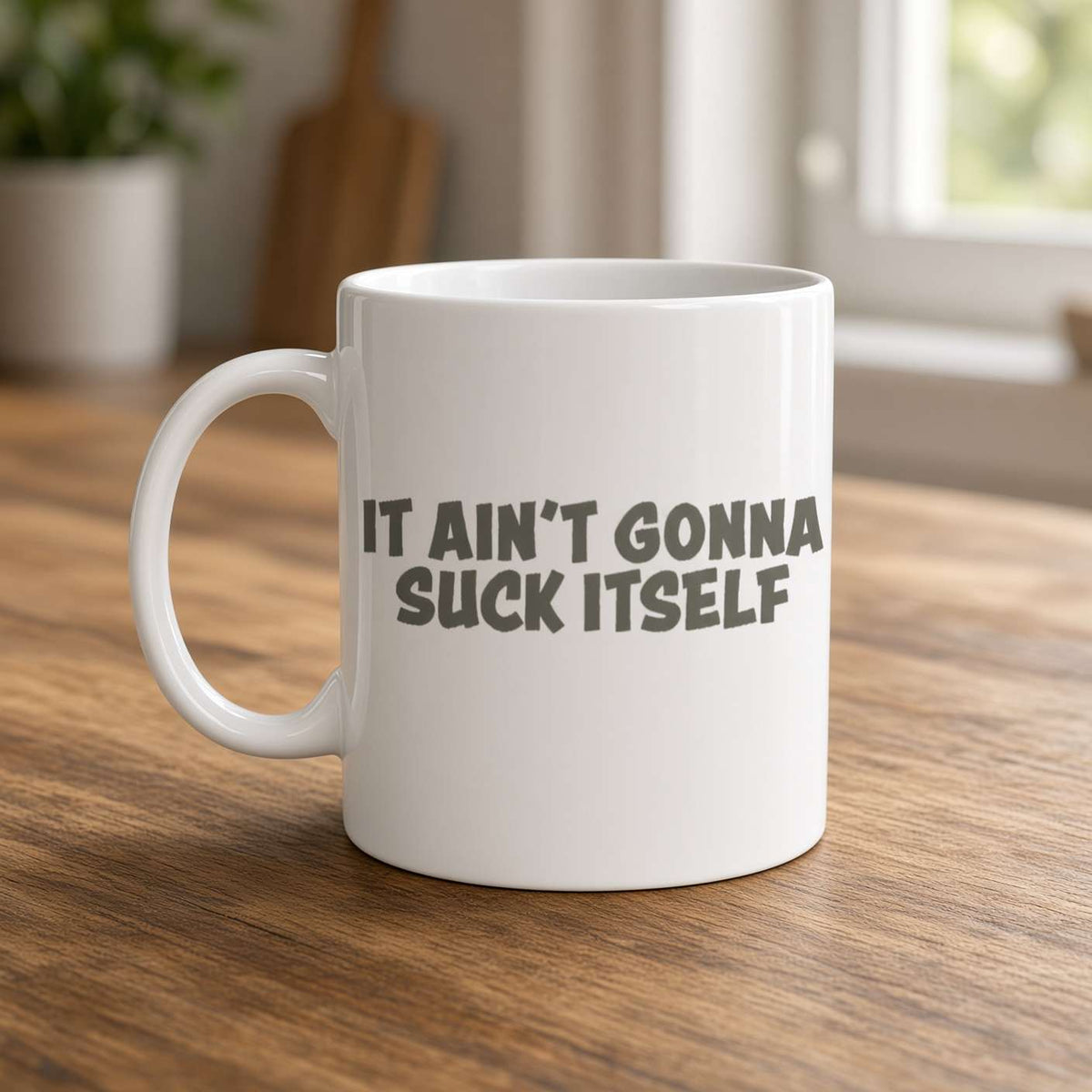 A white ceramic mug on a wooden table reads “It ain’t gonna suck itself,” with a softly lit kitchen background and a cheeky, humorous tone.