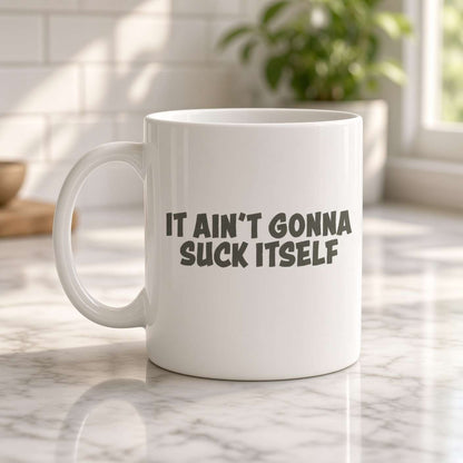 A white ceramic mug on a marble kitchen counter reads “IT AIN’T GONNA SUCK ITSELF.” Sunlight and a leafy plant create a bright, playful mood.