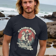 A man with long hair stands on a rocky beach, wearing a black funny graphic T-shirt with a humorous design of a Jesus, riding a unicorn. The text reads, 