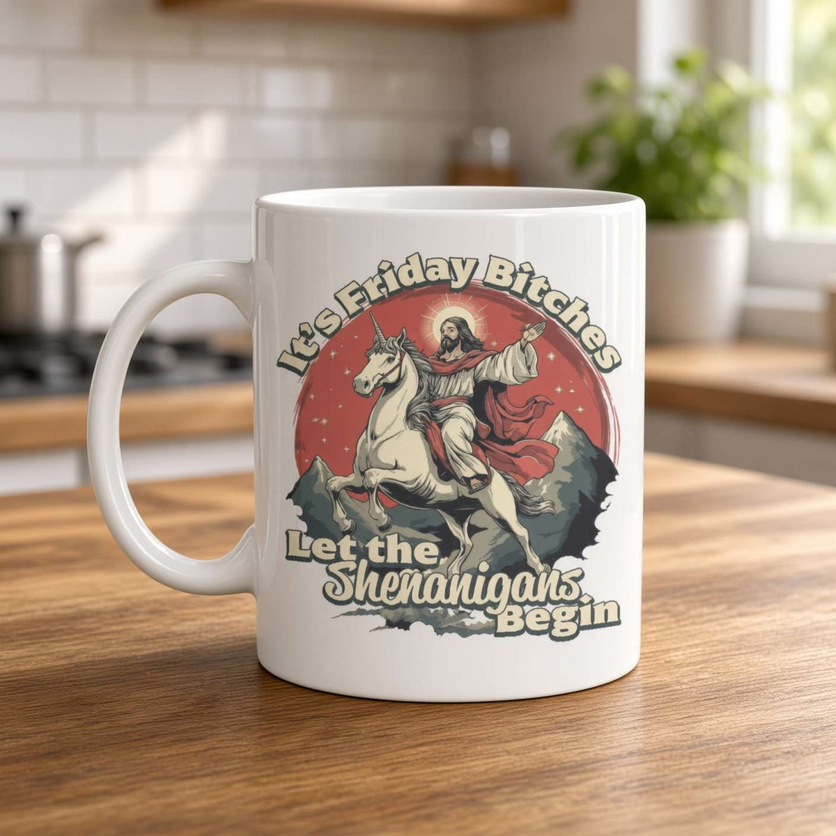 A white ceramic coffee mug with a humorous graphic print, sitting on a wooden table in a blurred kitchen setting. The graphic features a cartoon illustration of Jesus riding a unicorn in front of a red sun, with the text 'It's Friday Bitches Let the Shenangars Begin'.