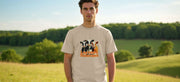 A man stands outdoors in a sunny field, wearing a beige funny T-shirt with cartoon cows and the text 