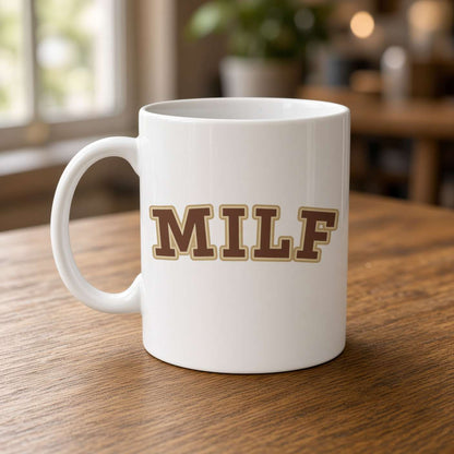 A white ceramic coffee mug with the word 'MILF' printed in brown block letters with a gold outline, sitting on a wooden table in a blurred indoor setting.