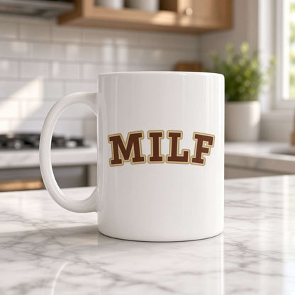 A white ceramic coffee mug with the word 'MILF' printed in brown varsity-style letters outlined in gold, sitting on a marble kitchen counter.