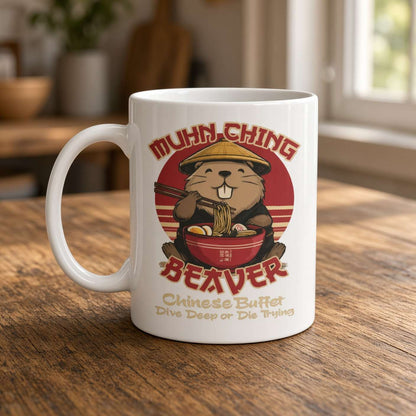 A white ceramic mug with a cartoon beaver eating ramen and the text 'MUHN CHING BEAVER' and 'Chinese Buffet Dive Deep or Die Trying', placed on a wooden table in a softly blurred indoor setting.