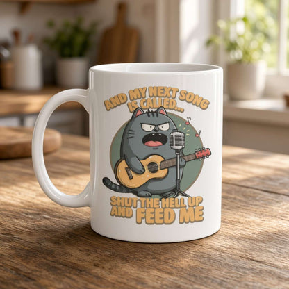 White coffee mug on a sunlit kitchen table, featuring a grumpy gray cat with a guitar and microphone. Text reads, “And my next song is called… Shut the hell up and feed me,” giving the design a humorous, irreverent tone.