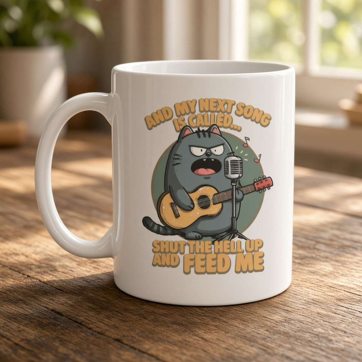 A white ceramic coffee mug with a cartoon cat playing guitar and the text 'AND MY NEXT SONG IS CALLED. SHUT THE HELL UP AND FEED ME', shown on a wooden table in a softly blurred indoor setting.