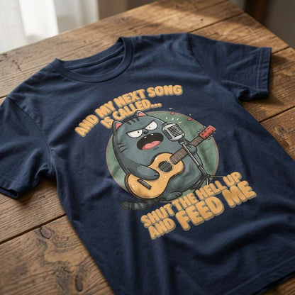 Navy blue t-shirt laid flat on a wooden table showing the full angry cat musician design and the witty “And My Next Song Is Called… Shut The Hell Up And Feed Me” text