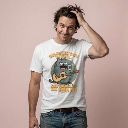 Man running a hand through his hair with a cheeky half-smile while wearing a white t-shirt featuring an angry cartoon cat on guitar singing “And My Next Song Is Called… Shut The Hell Up And Feed Me”