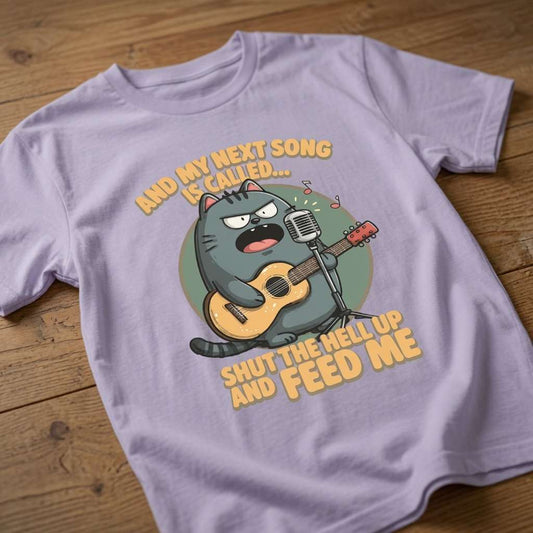 Lavender t-shirt laid flat on a wooden surface displaying the complete angry cat musician design and the relatable “And My Next Song Is Called… Shut The Hell Up And Feed Me” print
