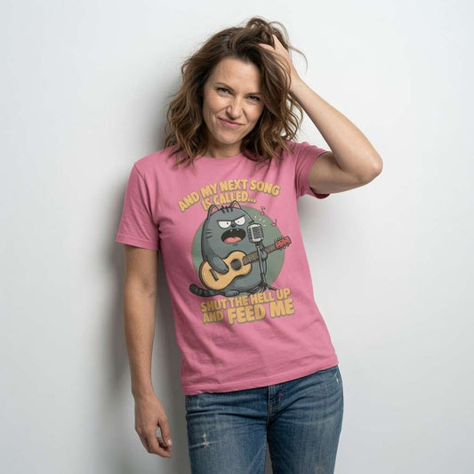 Woman with a playful half-smile and one hand in her hair wearing a pink t-shirt featuring the angry cartoon cat on guitar and the full “And My Next Song Is Called… Shut The Hell Up And Feed Me” slogan