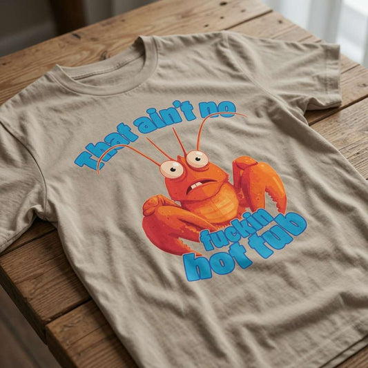 Beige t-shirt laid flat on a wooden table showing the full stressed lobster graphic and the rude “That ain’t no fuckin hot tub” text, classic witty animal tee