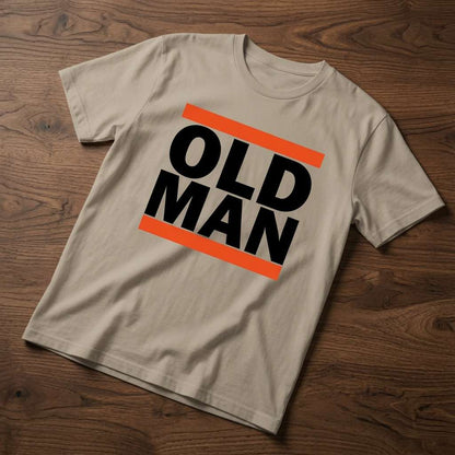Beige Old Man t-shirt laid flat on a wooden surface showing the bold black text framed by orange bars, simple and funny dad design