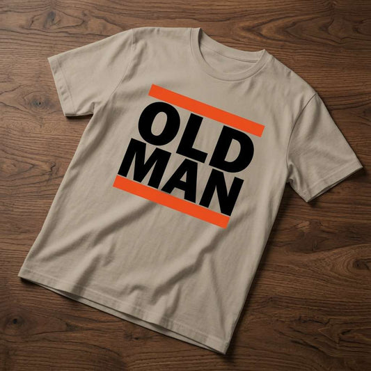 Beige Old Man t-shirt laid flat on a wooden surface showing the bold black text framed by orange bars, simple and funny dad design