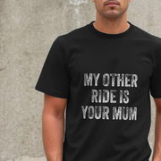 Person wearing a black funny slogan t-shirt with the offensive slogan 'My other ride is your mum' against a concrete wall.