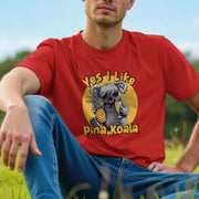 A man sits on grass wearing a red animal T-shirt with a playful koala graphic holding a pineapple and drink, captioned 