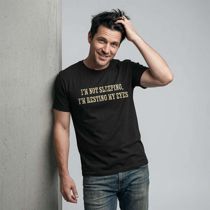 Man scratching his head with a knowing smile in a black I’m Not Sleeping, I’m Resting My Eyes t-shirt, funny mens witty slogan full of pure dad energy