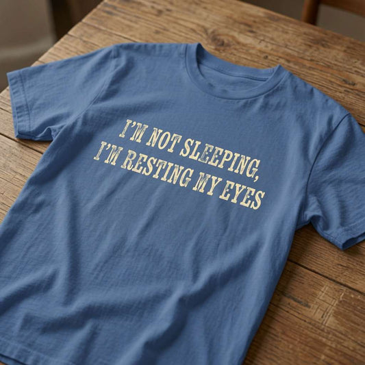 Bright blue I’m Not Sleeping, I’m Resting My Eyes t-shirt laid flat on a wooden surface with classic cream text, sleeping quote dad gift idea that every father will claim