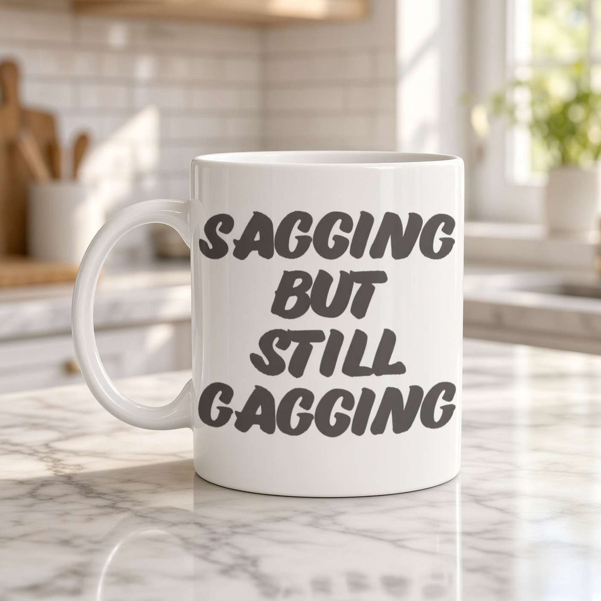 A white coffee mug on a sunlit marble kitchen counter reads “Sagging but still gagging” in bold lettering, conveying playful, cheeky humor.
