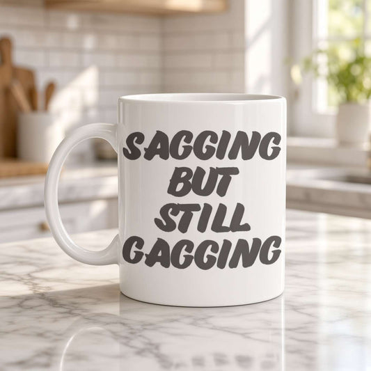 A white coffee mug on a sunlit marble kitchen counter reads “Sagging but still gagging” in bold lettering, conveying playful, cheeky humor.