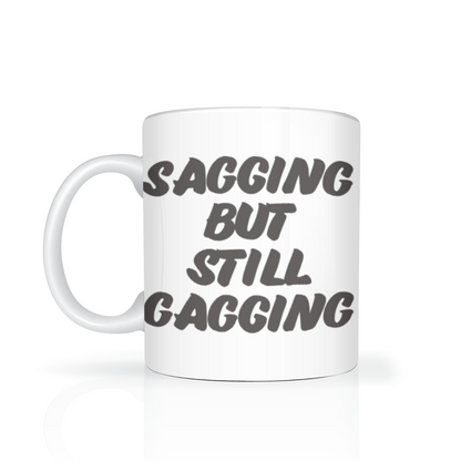 A white mug on a reflective surface reads “Sagging but still gagging” in bold dark lettering, with a playful, irreverent tone.