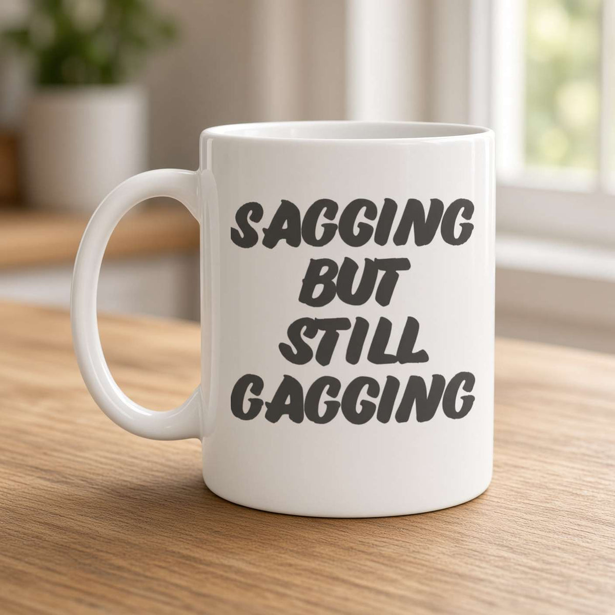 A white ceramic mug on a wooden table reads “Sagging but still gagging” in bold black lettering, with a bright, cozy kitchen blurred in the background.