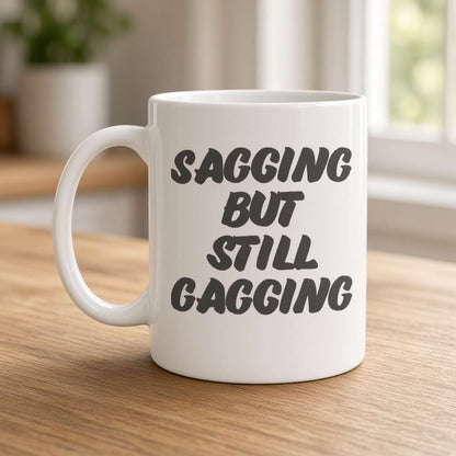 A white ceramic mug on a wooden table reads “Sagging but still gagging” in bold black lettering, with a bright, cozy kitchen blurred in the background.