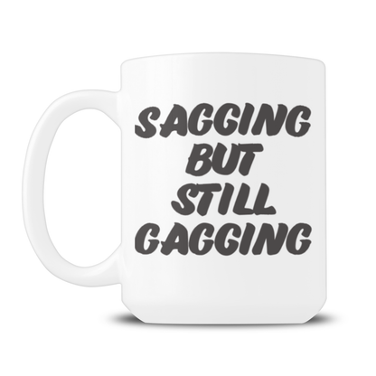 A white ceramic mug displays bold black lettering reading “Sagging but still gagging,” with a playful, cheeky tone against a plain white background.