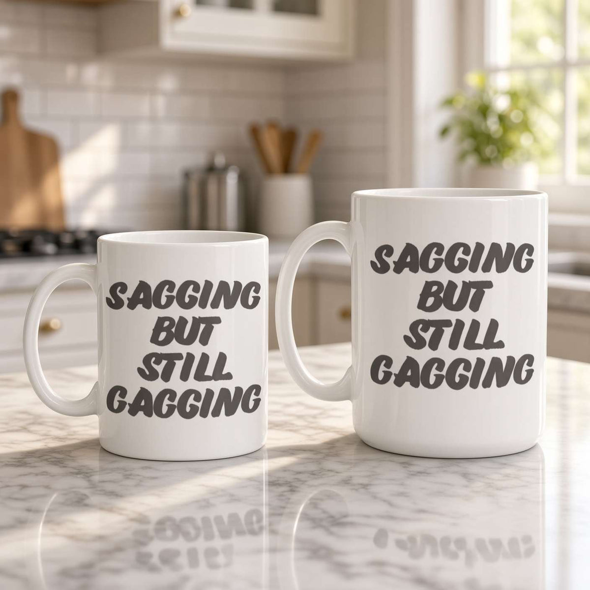 Two white ceramic mugs on a sunlit kitchen counter read “Sagging but still gagging,” with bold black lettering and reflections on the marble; playful, cheeky mood.