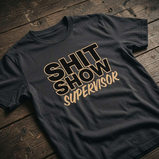 Black Shit Show Supervisor t-shirt laid flat on a wooden surface with bold outlined text, funny ladies relatable slogan design