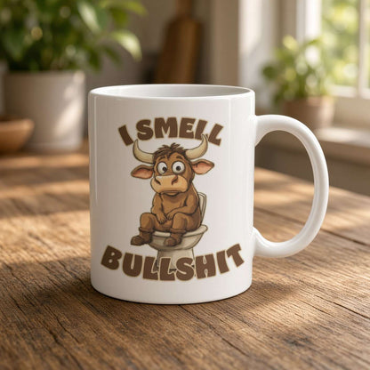 A white ceramic coffee mug with a cartoon bull sitting on a toilet and the text 'I SMELL BULLSHIT' printed on it, placed on a wooden table with a blurred indoor background.