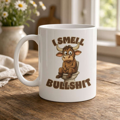 A white ceramic mug with a cartoon bull sitting on a toilet and the text 'I SMELL BULLSHIT' printed on the front, placed on a wooden table in a softly lit indoor setting.