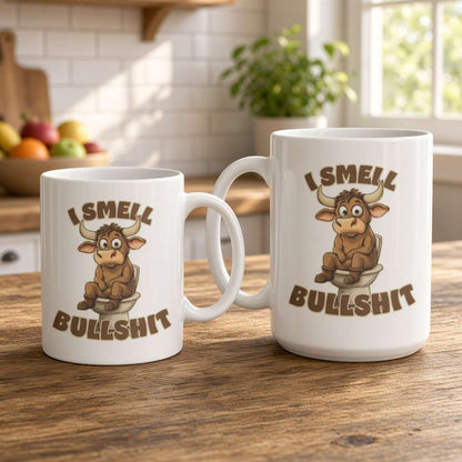 Two white ceramic mugs with a cartoon bull sitting on a toilet and the text 'I SMELL BULLSHIT' printed on them, displayed on a wooden table in a kitchen setting.