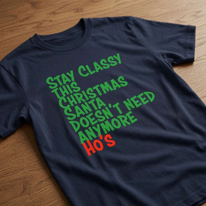 Navy Stay Classy This Christmas Santa Doesn’t Need Anymore Ho’s t-shirt laid flat on a wooden surface with green and red lettering, funny mens cheeky slogan xmas design